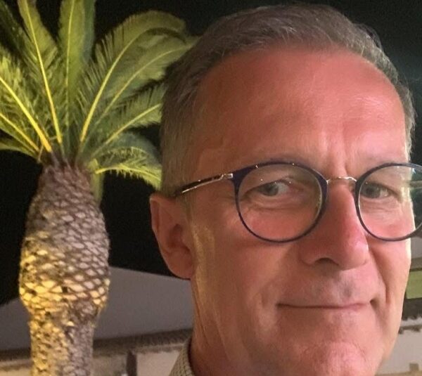 Uomo in elegante completo scuro con cravatta blu e occhiali, sorridente davanti a sfondo blu istituzionale