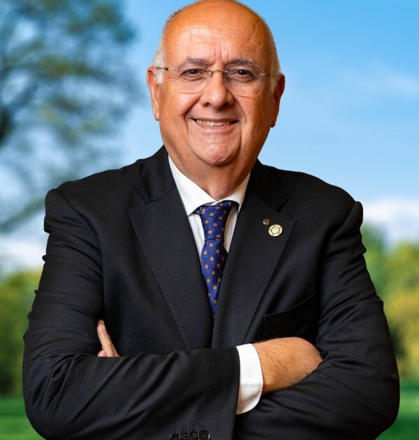 Uomo in giacca nera e cravatta blu con occhiali sorride con braccia incrociate su sfondo verde naturale