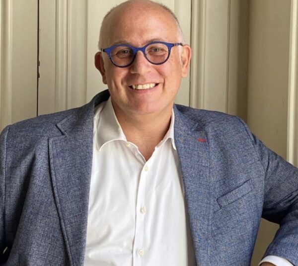 Uomo sorridente con occhiali blu e giacca grigia, camicia bianca, in posa professionale per associazione Rotarian Gourmets