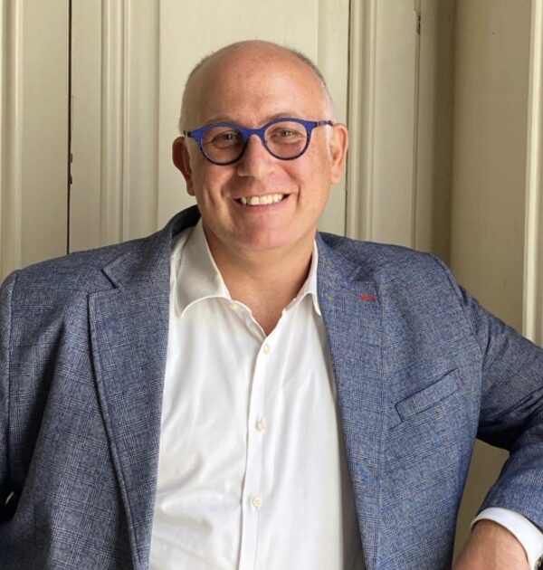 Uomo sorridente con occhiali blu e giacca grigia, camicia bianca, in posa professionale per associazione Rotarian Gourmets